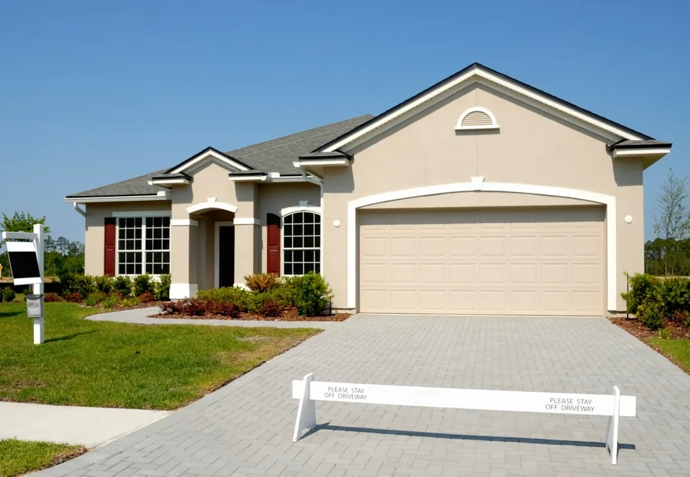 Beige stucco house with a tan double-wide raised-panel garage door over a paver driveway